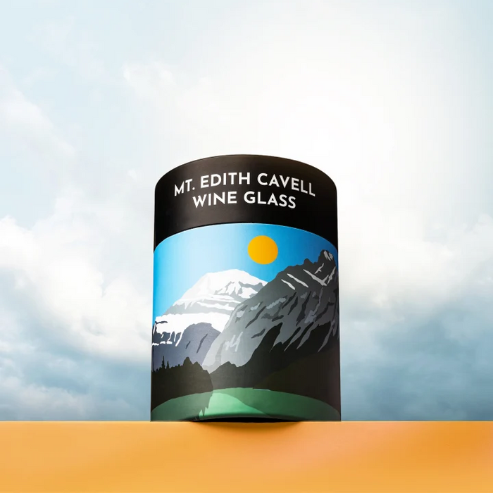 A cylindrical wine glass with a black top band reading 'MT. EDITH CAVELL WINE GLASS' and a mountain landscape label, shown standing on an orange surface against a cloudy sky background.