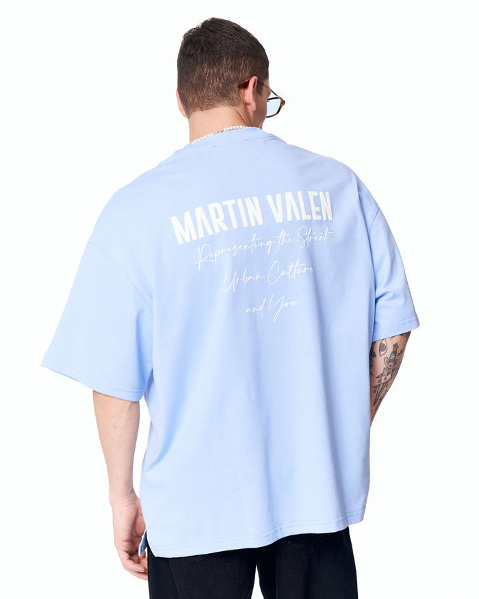 Martin Valen "Slogan" men's oversized blue T-shirt, rear three-quarter view worn with sunglasses, large white back print visible